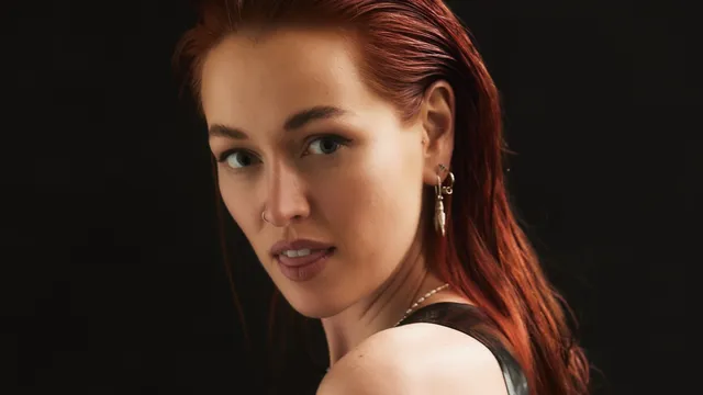 Foto van Bebe Bad die in de camera kijkt. Ze heeft rood haar, kleine tatoeages op haar armen en staat voor een zwarte achtergrond.