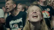 Blonde vrouw met neuspiercing schreeuwt met haar ogen dicht, op de achtergrond mensen staan gekleed in bandshirts.