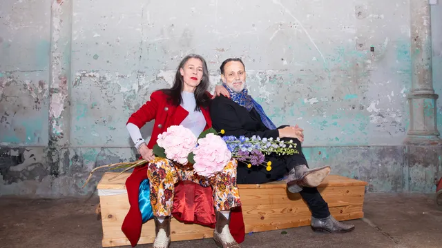 Adelheid Roosen en Hugo Borst zitten naast elkaar op een houten kist voor een verweerde muur met afbladderende verf. Adelheid draagt een rood jasje, gebloemde broek en witte coltrui en houdt een bos roze hortensia's vast; Hugo draagt een zwart jack met een blauwe sjaal en houdt blauwe en gele bloemen vast. Ze kijken rustig en direct in de camera.
