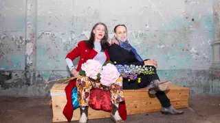 Adelheid Roosen en Hugo Borst zitten naast elkaar op een houten kist voor een verweerde muur met afbladderende verf. Adelheid draagt een rood jasje, gebloemde broek en witte coltrui en houdt een bos roze hortensia's vast; Hugo draagt een zwart jack met een blauwe sjaal en houdt blauwe en gele bloemen vast. Ze kijken rustig en direct in de camera.