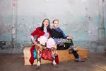 Adelheid Roosen en Hugo Borst zitten naast elkaar op een houten kist voor een verweerde muur met afbladderende verf. Adelheid draagt een rood jasje, gebloemde broek en witte coltrui en houdt een bos roze hortensia's vast; Hugo draagt een zwart jack met een blauwe sjaal en houdt blauwe en gele bloemen vast. Ze kijken rustig en direct in de camera.