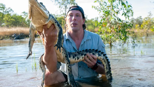 Freek Vonk staat in het water en houdt een krokodil vast in zijn handen. De kop van de krokodil is naar de camera toe gericht.