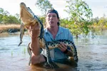Freek Vonk staat in het water en houdt een krokodil vast in zijn handen. De kop van de krokodil is naar de camera toe gericht.