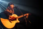 Een muzikant met een zwarte muts zingt in een microfoon en speelt op een akoestische gitaar op een donker podium met sfeervolle podiumverlichting.