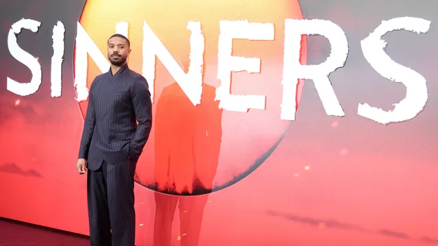 Acteur Michael B Jordan, gekleed in een zwart krijtstreetpak, staat voor een muur waarom het woord Sinners te lezen is.