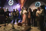 Foto van een bijeenkomst of tentoonstelling in een kerkinterieur. Centraal staat een kunstwerk op een laag platform gevuld met grijs zand, waarin een grote plant met donkergroene nerven en een cirkelvormig rood paneel staan. Op de achtergrond staat een gemêleerd publiek onder de verlichte gewelven en ronde roosvensters van de kerk. De sfeer is informeel en sociaal.