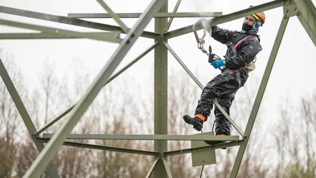 Een foto van een man in een waterdicht pak die in een hoogspanningsmast staat met een hogedrukspuit om hem schoon te maken. Het is een grijze dag.