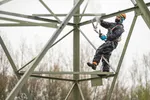 Een foto van een man in een waterdicht pak die in een hoogspanningsmast staat met een hogedrukspuit om hem schoon te maken. Het is een grijze dag.