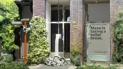 Een digitale illustratie van een bakstenen gevel, deels overwoekerd door groene planten. Links staat een beveiligingscamera op een oranje paal. In het midden hangt een lange strook papier aan een ketting, met onderaan een hoop opgerold papier. Rechts hangt een poster met de tekst ‘Mees is taking a toilet break. 5 seconds ago’.