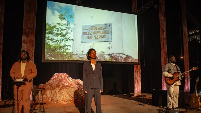 Voorstelling beeld met 3 mensen die voor een digitaal scherm staan met op dat scherm een plakkaat van surinaamse oorlogvrijwilligers