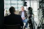 Een man zit in een interviewopstelling in een modern kantoor met grote ramen. Op de voorgrond zijn camera's en de rug van een interviewer zichtbaar.