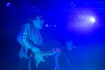 Een dynamisch actieshot van een zanger/gitarist die vol overgave schreeuwt in een microfoon terwijl hij gitaar speelt. Hij draagt een zonnebril en een blauw sportjack met witte strepen op de mouwen. Het podium is gehuld in een intens blauw licht, waarbij op de achtergrond een tweede bandlid zingend zichtbaar is.