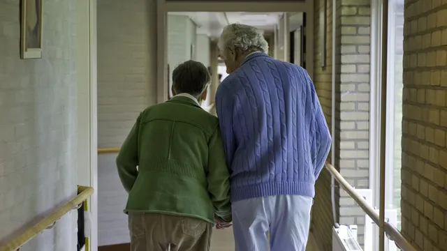 Twee oudere mensen op de rug gefotografeerd. Ze lopen hand in hand door een gang in het verpleeghuis.