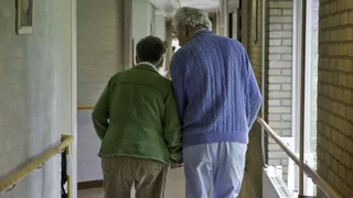 Twee oudere mensen op de rug gefotografeerd. Ze lopen hand in hand door een gang in het verpleeghuis.