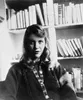 Een oude zwart-wit foto van de dichter Sylvia Plath. Ze zit voor een boekenkast en kijkt met een kleine glimlach de camera in.