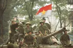 Groep Vietnamese soldaten in groene uniformen en helmen zit en staat op een tank, met een rood-blauwe vlag met gele ster wapperend boven hen. Ze lachen, kijken geconcentreerd of poseren. De scène speelt zich buiten af, omringd door bomen.