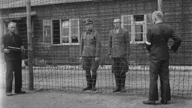 2 personen, SS-kampbewaker B.J. Westerveld (rechts) en kampcommandant K.P. Berg, staan achter een hek. Berg draagt een uniform. Ze worden bewaakt door twee mannen van de Binnenlandse Strijdkrachten.