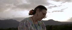 Persoon met kleurrijke blouse staat buiten in een bergachtig landschap, met bewolkte lucht op de achtergrond.