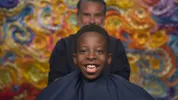 Jehneo zit in de kappersstoel, hij lacht met open mond en kijkt in de camera.