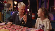 Presentator Maarten Bel knutselt met een kind. Hij heeft zijn vinger in de lucht. Ze kijken elkaar aan.