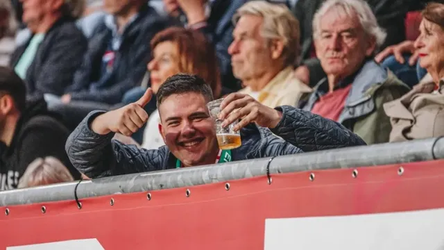 Een jongen op een tribune steekt vrolijk zijn duim op naar de camera terwijl hij een biertje vasthoudt.