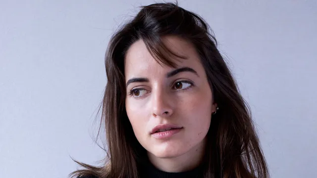 Vrouw met bruin haar tegen lichtblauwe achtergrond. Ze draagt een zwarte trui en kijkt naar links.
