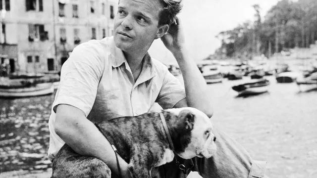 American journalist and writer Truman Capote sitting on a low wall in front of the small harbour of Portofino with his bulldog (Charlie J. Fatburger) in his arm. Portofino, 1950s