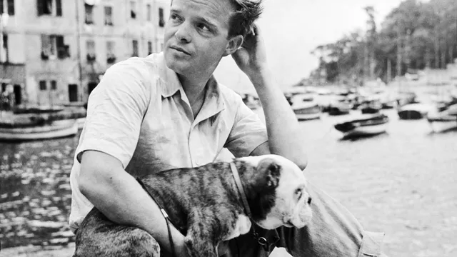 American journalist and writer Truman Capote sitting on a low wall in front of the small harbour of Portofino with his bulldog (Charlie J. Fatburger) in his arm. Portofino, 1950s