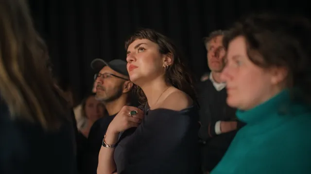 Een groep mensen staat aandachtig te kijken in een donkere ruimte, vermoedelijk een theatervoorstelling of tentoonstelling. Op de voorgrond staat een jonge vrouw met donker golvend haar; ze kijkt omhoog met een geconcentreerde, licht ontroerde blik en houdt haar hand aan haar keel. Om haar heen zijn meerdere toeschouwers zichtbaar, waaronder een man met een pet en bril en een vrouw in een turquoise trui. De sfeer is intiem en geladen.