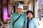 De ouders van Pete staan in een winkelstraat. Ze kijken met een lach in de camera. Er zijn Chinese winkels, etalages en reclame borden te zien.