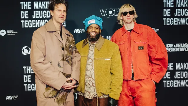 Drie mannen (van De Jeugd van Tegenwoordig) poseren voor een VPRO-fotowand: links een witte man in een beige jas, in het midden een zwarte man met een blauwe muts en groene jas die glimlacht, rechts een witte man met blond haar, zonnebril en een feloranje outfit.