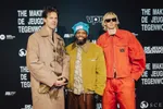 Drie mannen (van De Jeugd van Tegenwoordig) poseren voor een VPRO-fotowand: links een witte man in een beige jas, in het midden een zwarte man met een blauwe muts en groene jas die glimlacht, rechts een witte man met blond haar, zonnebril en een feloranje outfit.