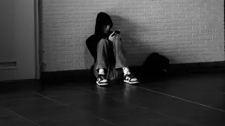Zwart-wit foto van een middelbare scholier die in de gang tegen de muur zit met zijn telefoon en een zwarte capuchon draagt.
