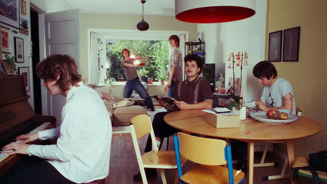 De bandleden zijn allemaal op een andere manier gepositioneerd in de woonkamer. Een iemand achter de piano, twee aan tafel en twee voor het raam