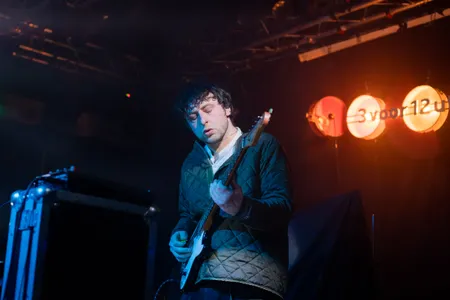 Een muzikant met donker, warrig haar speelt geconcentreerd op een elektrische gitaar tijdens een live-optreden. Hij draagt een gewatteerde groene jas over een wit hemd. Op de donkere achtergrond branden drie ronde, oranje lampen met het logo van "3voor12 Utrecht". Links op de voorgrond is een zwarte flightcase zichtbaar.