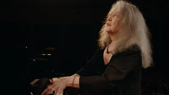 Een dame met grijze krullen speelt piano tegen een zwarte achtergrond.