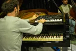Een muzikant van achteren gezien die een compacte Korg synthesizer bespeelt, die bovenop een zwarte vleugelpiano is geplaatst. Zijn linkerhand draait aan een knop terwijl zijn rechterhand de toetsen van de synthesizer indrukt. Op de achtergrond is een tweede muzikant zichtbaar met een contrabas in een studio-achtige setting met houten panelen en muziekstandaarden.