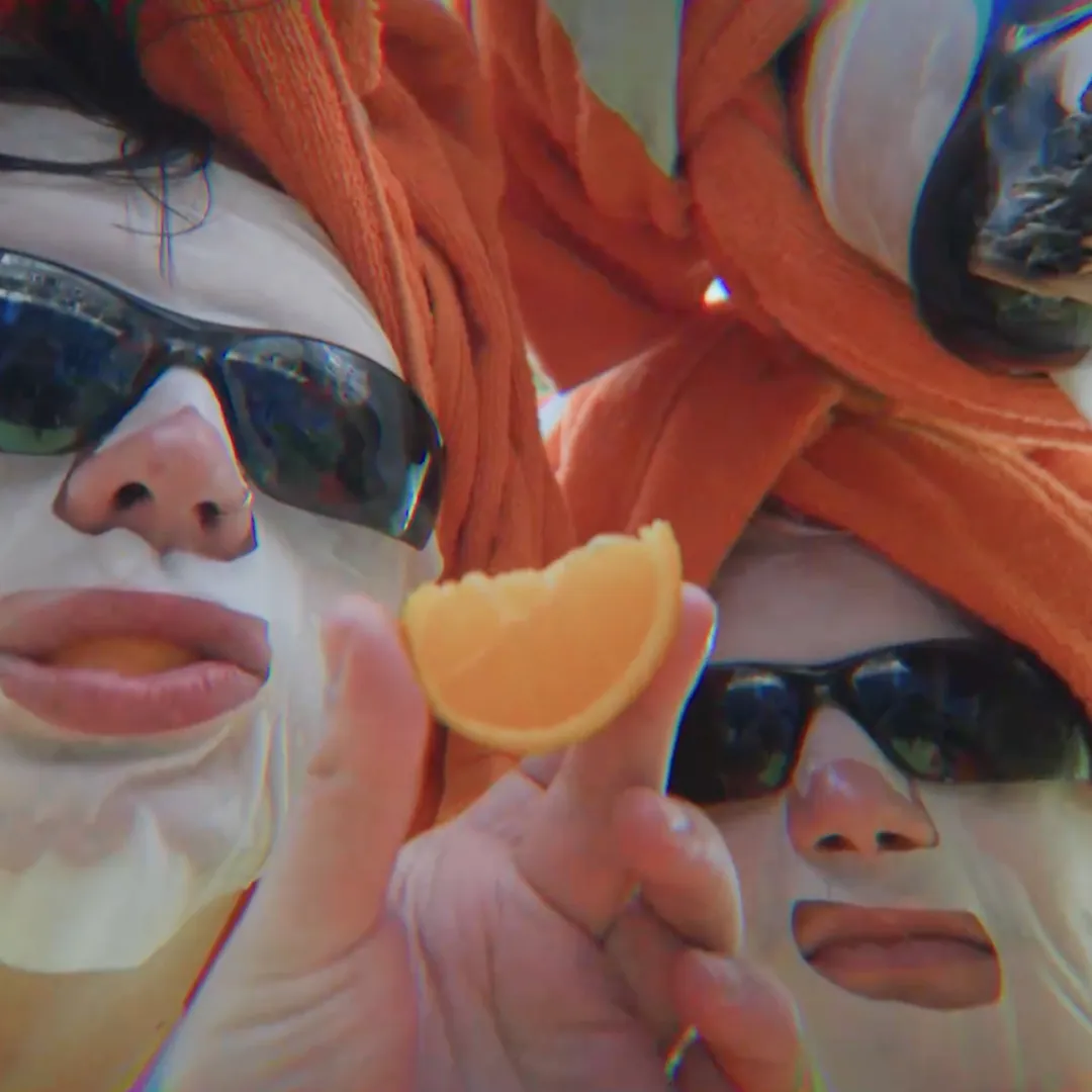 Drie mensen die in een close up foto van hun gezicht maken met dezelfde zonnebril, gezichtsmasker en oranje handoek up hun hoofd, terwijl een van hun een stuk sinasappel in hun hand heeft.