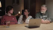 Een tienerjongen zit aan de keukentafel met zijn vader en moeder achter een laptop te praten.