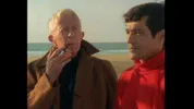 Twee mannen staan naast elkaar op een strand met de zee op de achtergrond. De man links draagt een bruine jas en houdt een pijp tegen zijn mond, terwijl de man rechts een opvallende rode coltrui draagt en voor zich uit kijkt.