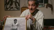 Een oudere man zit binnen aan tafel en houdt een ingelijste zwart-witfoto vast van een jonge, gespierde man die in het water staat.