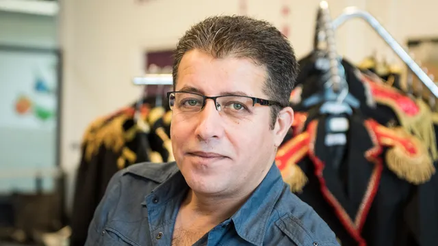 Portret van Bilal, een man met donker haar en een zwarte bril in een grijs overhemd. Hij kijkt rustig in de camera. Op de achtergrond hangen zwarte kostuums met goud en rood borduurwerk aan een kledingrek, vermoedelijk in een theater of kostumerie.