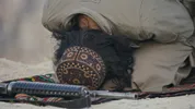 Man in grijs gewaad met kufi op zijn hoofd knielt, voor hem ligt een wapen.
