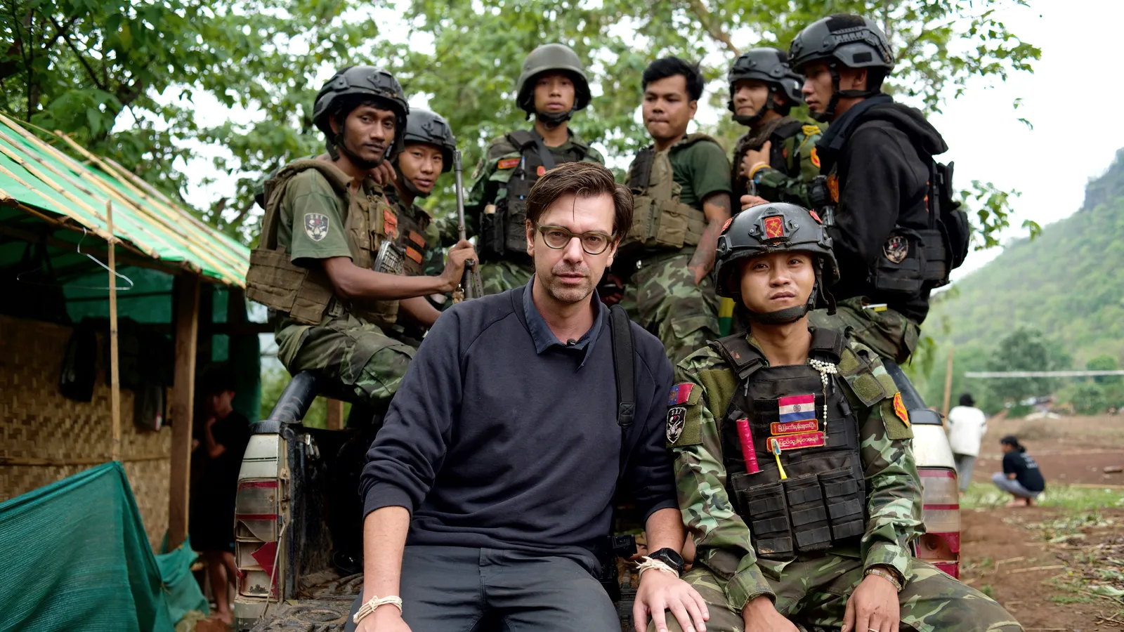 Een ernstig kijkende witte man met bril, Ruben Terlou, zit op de achterkant van een pick-up truck naast een gewapende Aziatische man in camouflage-uniform en helm. Achter hen zitten en staan nog zes gewapende Aziatische mannen in militaire uitrusting. Op de achtergrond bomen, een bamboehut en een heuvelachtig landschap.
