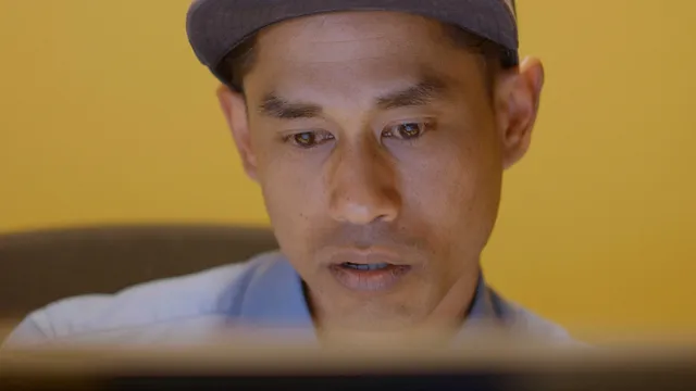 Eko zit achter zijn computer, hij kijkt geschrokken. Het scherm zie je alleen van de achterkant. Eko heeft een pet op en heeft bruine ogen.