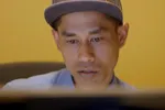 Eko zit achter zijn computer, hij kijkt geschrokken. Het scherm zie je alleen van de achterkant. Eko heeft een pet op en heeft bruine ogen.