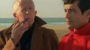 Een oudere witte man in bruine jas rookt op het strand, naast een jongere man met zwart haar in rode coltrui die opzij kijkt; de zee ligt op de achtergrond.