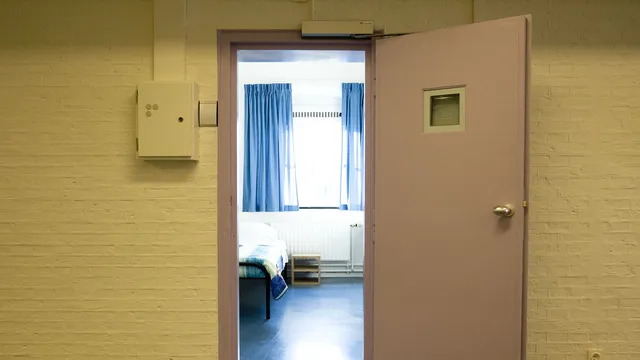 Een roze celdeur staat open, daarachter zie je een bed in een sober ingerichte kamer met blauwe vloer en blauwe gordijnen.