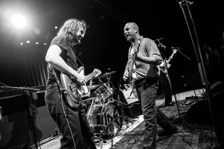 Foto van de gitarist en de bassist van Iguana Death Cult die tegenover elkaar staan en naar elkaar glimlachen tijdens het spelen.