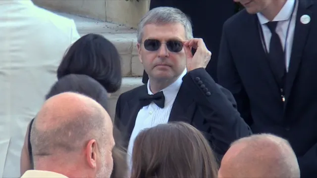 Een witte man met grijs haar in smoking en zwarte zonnebril kijkt recht de camera in terwijl hij tussen andere gasten zijn bril aanraakt.